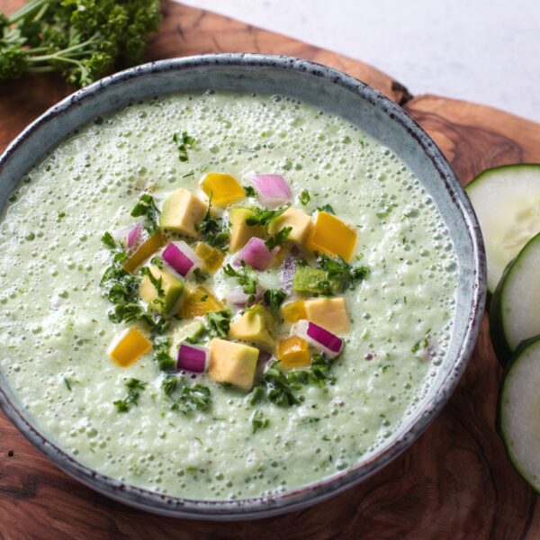 Frothy light green goddess cold soup topped with apples, red onion and parsley, served with cucumbers on a raw-edge wood cutting board