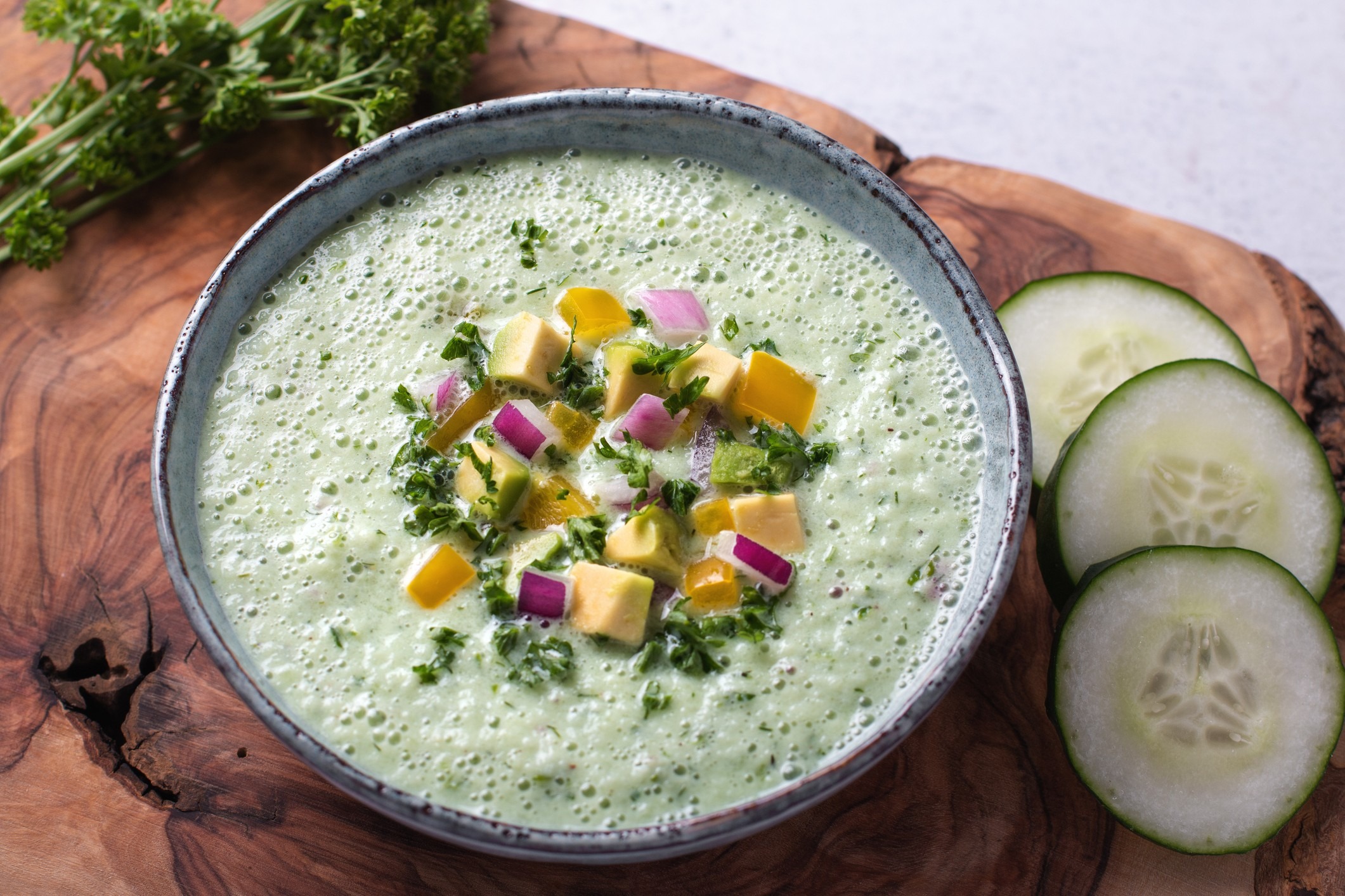 Frothy light green goddess cold soup topped with apples, red onion and parsley, served with cucumbers on a raw-edge wood cutting board