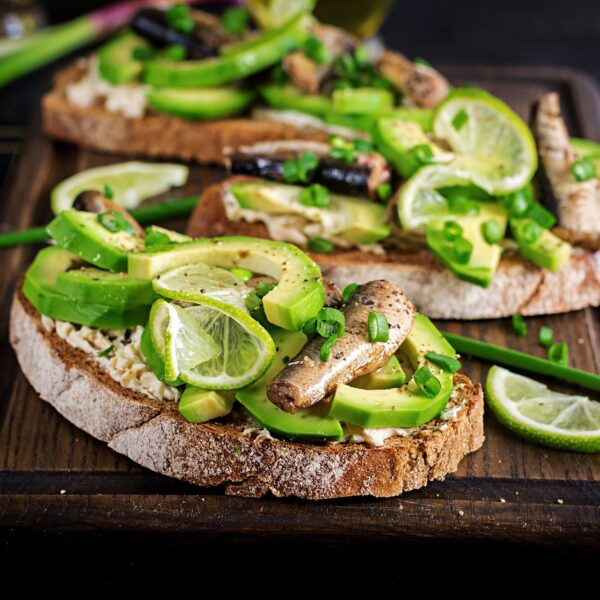 Three open-faced sandwiches of toasted artisan bread with avocado and smoked mackerel. Garnished with green onions and lime slices. On a wooden cutting board.
