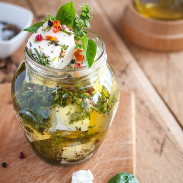 Glass jar filled with pickled chucks of white feta, red peppercorns and herbs on a weathered cutting board