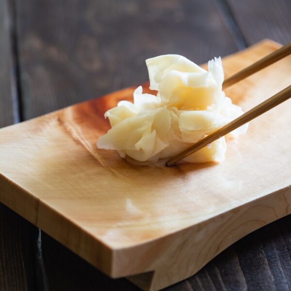 A bite of pickled ginger held with chopsticks over a traditional Japanese platter on a dark brown wood table.