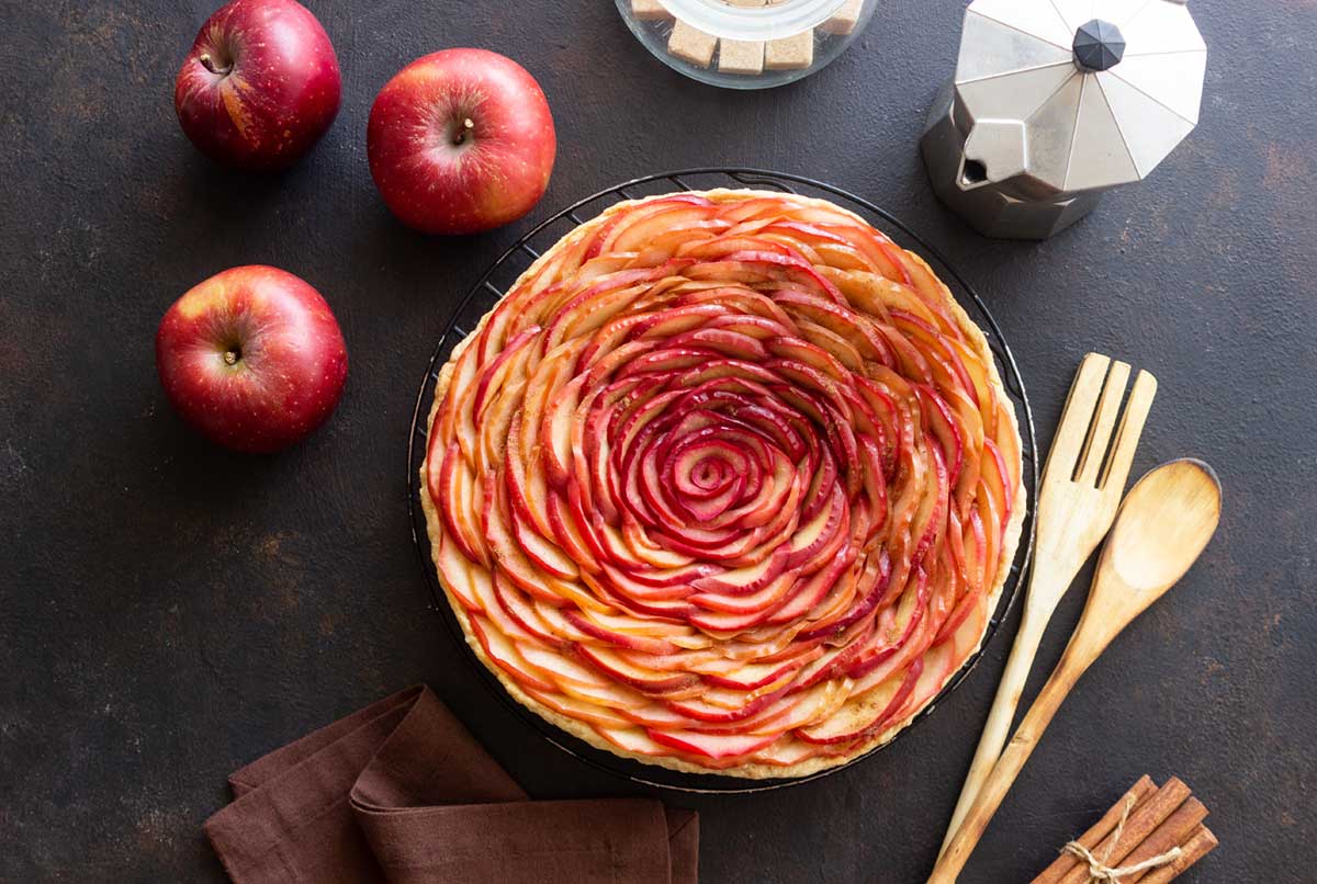 elegant apple rose tart with apples and baking utensils on a dark background, top view.