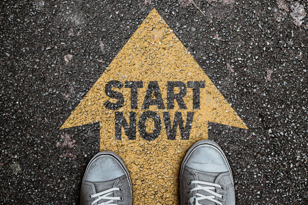high angle view of person standing on arrow marking on asphalt, road with text START NOW, motivation concept